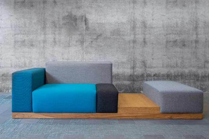 Modular sectional sofa in teal, blue, and gray tones on a wooden base, set against a concrete wall, modern minimalist mood
