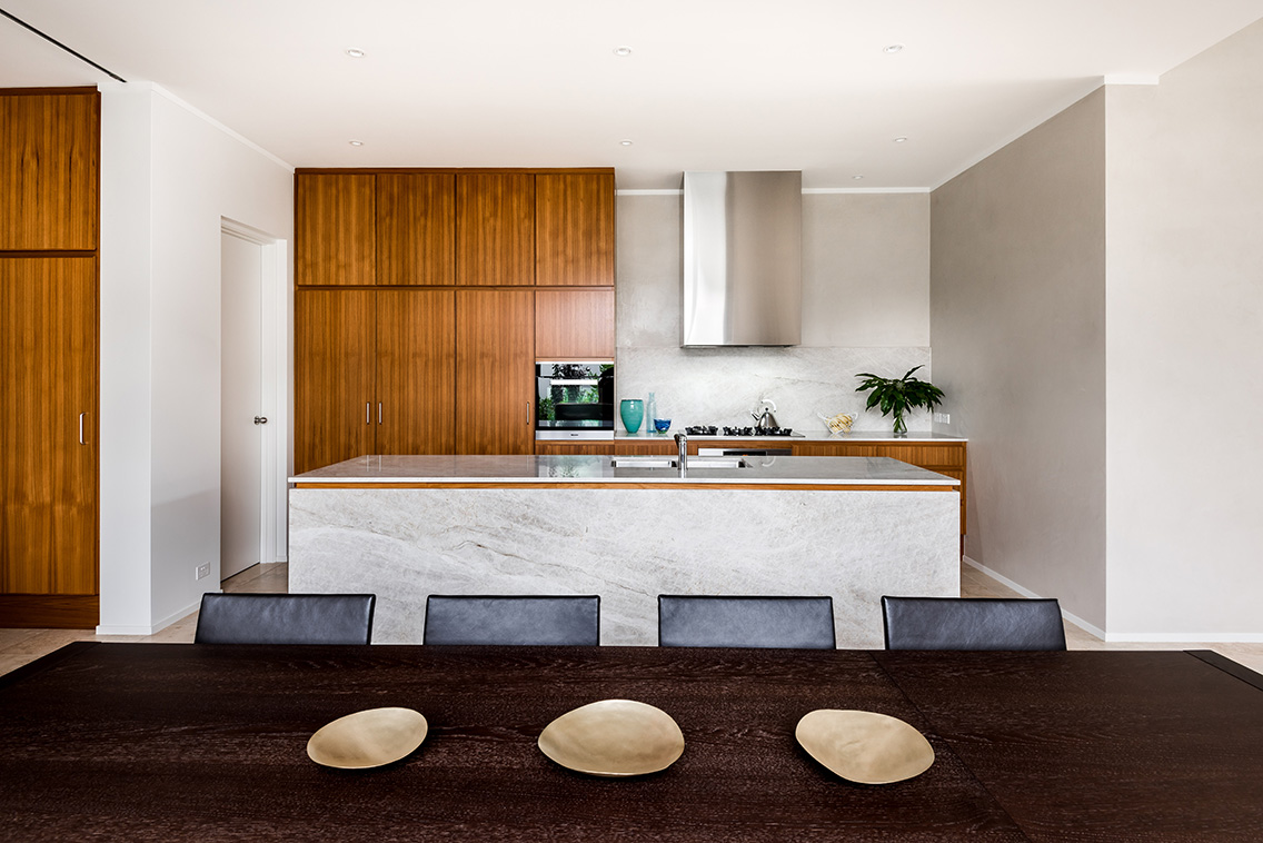 Modern kitchen with wooden cabinetry, marble island, stainless range hood, and dark dining table with decorative plates