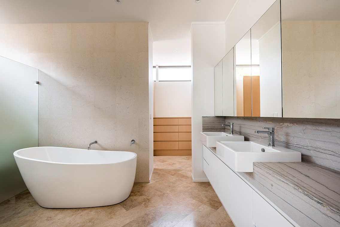 Modern bathroom with freestanding oval white tub, double vanity with marble backsplash, large mirrors, and stone-tiled floor