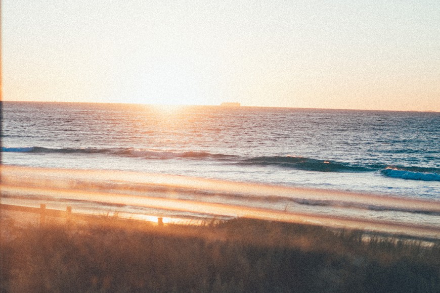 Ocean sunset with waves rolling onto shore, viewed through dune grass in foreground, warm orange and blue tones, calm reflective mood