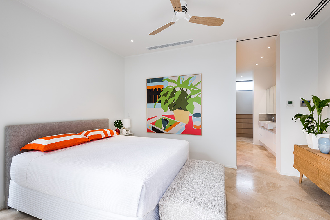 Bright bedroom with white bed, orange and white striped pillows, colorful wall art, ceiling fan, and wooden dresser; clean and airy